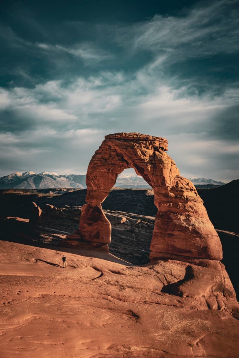 Delicate Arch with a hiker for scale and snow-capped mountains under dramatic clouds