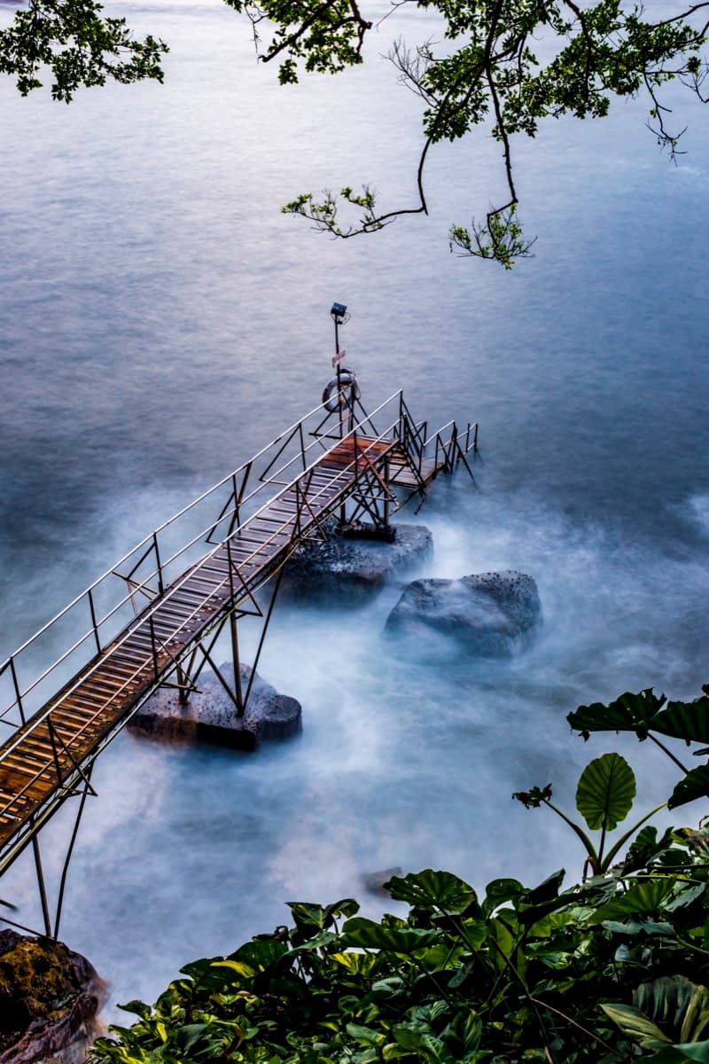 Long-exposure shot of a metal footbridge extending over misty rocks into calm sea water