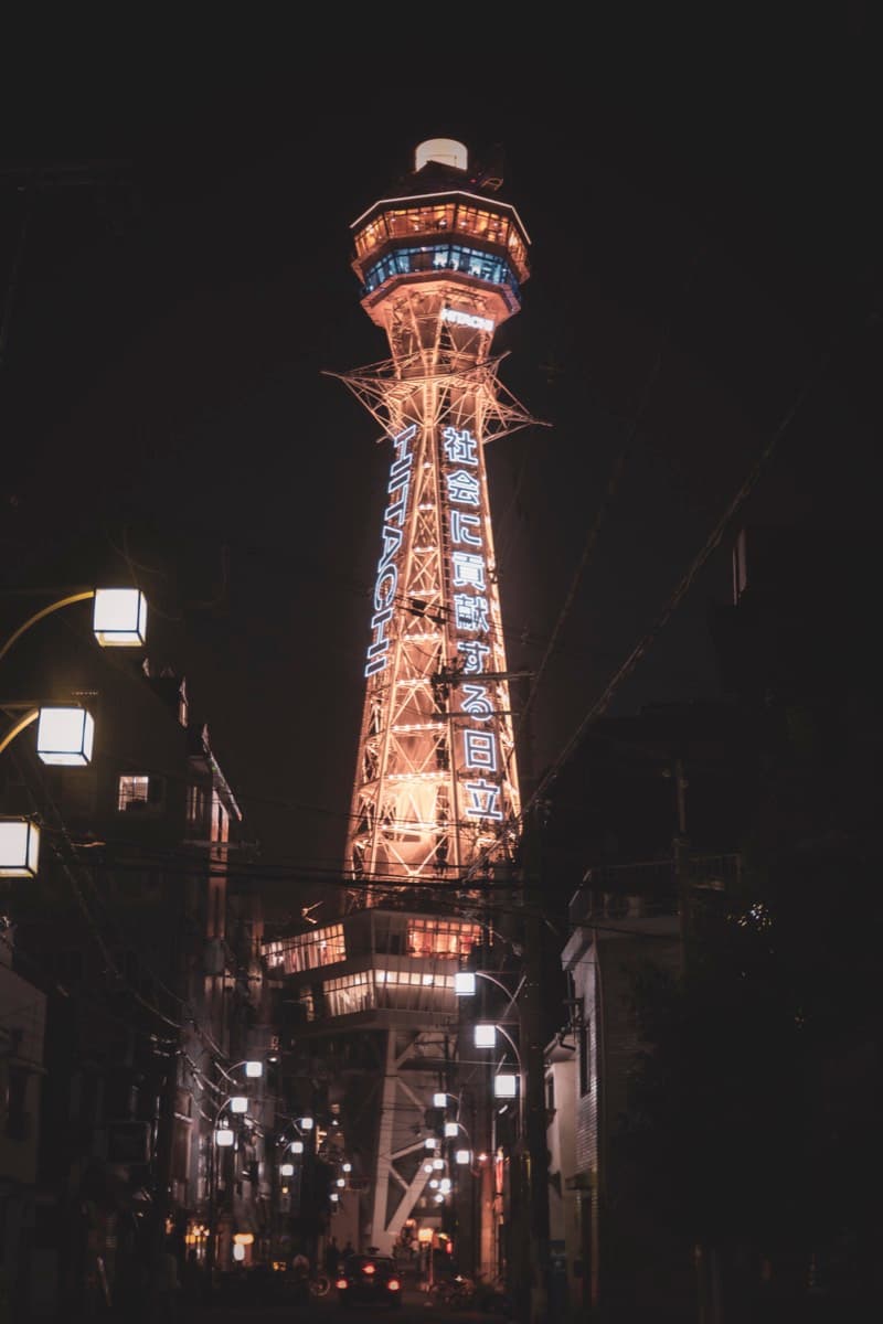 Tsutenkaku Tower illuminated at night with neon signage viewed from a narrow lantern-lined street